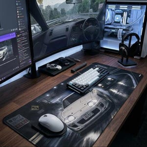 May include: A computer desk setup featuring a large desk mat with a white sports car graphic, a white wireless mouse, a mechanical keyboard with white and black keys, a monitor displaying a car interior, and a PC tower with internal lighting.