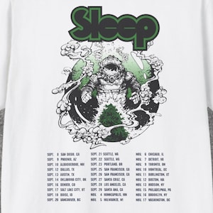 Op de afbeelding: Een wit T-shirt met korte mouwen en een afbeelding op de achterkant. Het ontwerp bevat het woord Sleep in het groen, een illustratie van een astronaut en een lijst met tourdata en steden van 8 september tot 17 november in zwarte tekst.