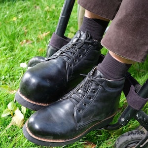 May include: A pair of black leather lace-up boots with a rounded toe, thick black soles, and black laces. The boots are worn with dark brown trousers and black socks, positioned on the footrests of a black metal wheelchair frame.