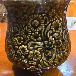 May include: A dark brown ceramic planter with a glossy finish and a raised, embossed floral pattern covering the exterior. The vessel features a wide, flared rim with a lighter tan glaze and stands on three small, integrated feet at the base.