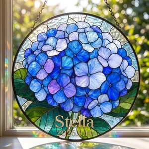 以下が含まれることがあります： 青と紫の紫陽花の花と緑の葉が描かれた円形のステンドグラスのサンキャッチャー。金色の文字で「Stella」という名前と「2026.9.14」という日付が記されており、金属製のチェーンで窓際に吊り下げられています。