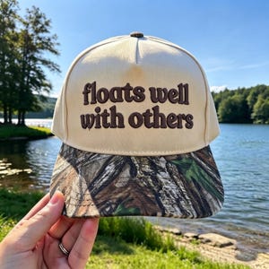 May include: A cream-colored baseball cap with a brown embroidered text that reads "floats well with others" on the front panel. The cap features a camouflage pattern on the brim and a structured crown with a small brown button on top.