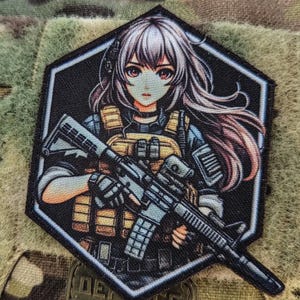Puede incluir: Un parche hexagonal bordado que muestra un personaje de estilo anime con cabello largo y claro, vistiendo un chaleco táctico color canela sobre una camisa oscura. El personaje sostiene un rifle negro con ambas manos. El diseño tiene un borde negro.