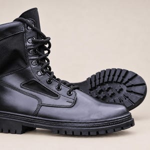 Puede incluir: Una bota táctica negra con una sección inferior de cuero liso, un cuello de tela alto y una suela de goma gruesa con tacos. La bota incluye cordones negros pasados por ojales metálicos y un patrón de rodadura visible en el talón.