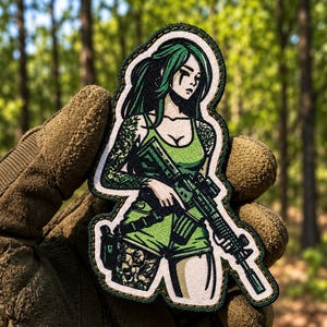Op de afbeelding: Een stoffen patch met een geïllustreerd personage met lang groen haar, gekleed in een groen hemdje en korte broek, die een zwart geweer vasthoudt. Het personage heeft zichtbare tatoeages op armen en benen en een donkergroene gestikte rand.