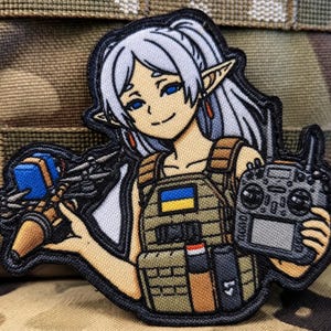 May include: An embroidered patch featuring an anime-style elf character with long white hair, wearing a tan tactical vest with a blue and yellow flag patch. The character holds a small drone in one hand and a remote control transmitter in the other.