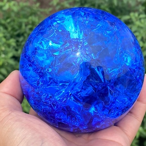 Könnte beinhalten: Ein kugelförmiges Objekt aus leuchtend blauem Glas mit einer ausgeprägten, rissigen Textur auf der gesamten Oberfläche. Das Innere der Kugel zeigt ein komplexes Netzwerk aus Brüchen, die das Licht einfangen und ein facettiertes Aussehen erzeugen.
