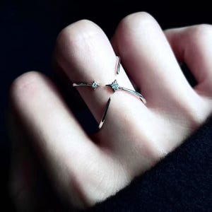 May include: A silver-toned open ring featuring a cross-over design with two pointed star-shaped accents. Each star-shaped point is set with a small, clear, sparkling stone at its center. The band is thin and polished, wrapping around the finger.