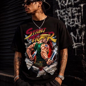 May include: A black short-sleeve t-shirt featuring a graphic print of the Street Fighter character Guile. The design shows the character in a green tank top holding two combat knives, framed by a torn metal effect with the Street Fighter logo.