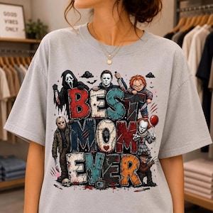 May include: A light gray t-shirt featuring a graphic print with the text BEST MOM EVER in large, distressed, multi-colored block letters. The design includes illustrations of five horror movie characters surrounding the text with knives and a balloon.