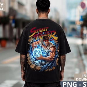 Puede incluir: Una camiseta negra de manga corta con un estampado gráfico en la espalda. El diseño muestra el logotipo de Street Fighter sobre la ilustración de un personaje musculoso con un gi blanco, sosteniendo una esfera de energía azul brillante ante un muro de piedra.