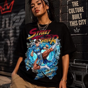 May include: A black short-sleeve t-shirt featuring a graphic print of the Street Fighter logo and the character Chun-Li in a dynamic kicking pose. The design is set against a blue swirling energy background with a torn paper effect on the fabric.