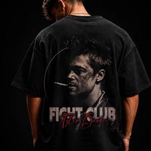 Può includere: Una t-shirt nera a maniche corte con una stampa grafica sul retro. Il disegno mostra il ritratto di un uomo con una sigaretta, il testo "FIGHT CLUB" in un carattere usurato e il nome "Tyler Durden" in corsivo rosso.