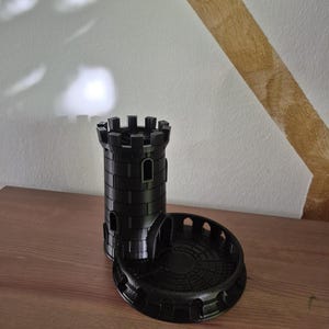 Puede incluir: Una torre de dados negra impresa en 3D que presenta una torreta de castillo cilíndrica con almenas y ventanas arqueadas, conectada a una base circular con un muro bajo y un suelo texturizado diseñado para atrapar y contener los dados.