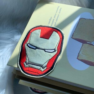 Puede incluir: Un parche de tela bordado que presenta el ic&oacute;nico casco rojo y dorado de Iron Man. El dise&ntilde;o muestra una m&aacute;scara estilizada con contornos negros, una secci&oacute;n central en la frente y ranuras estrechas para los ojos en hilo vibrante.