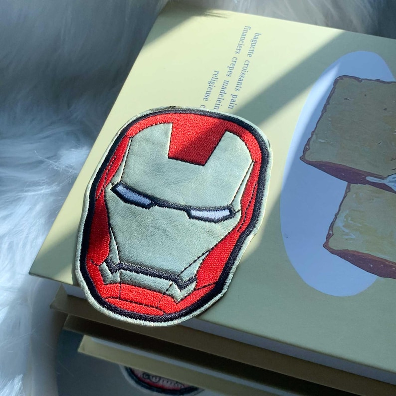 Puede incluir: Un parche de tela bordado que presenta el ic&oacute;nico casco rojo y dorado de Iron Man. El dise&ntilde;o muestra una m&aacute;scara estilizada con contornos negros, una secci&oacute;n central en la frente y ranuras estrechas para los ojos en hilo vibrante.