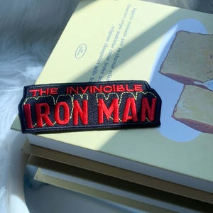Puede incluir: Un parche bordado rectangular con fondo negro y letras rojas que dicen THE INVINCIBLE IRON MAN. El texto est&aacute; delineado con costuras amarillas y el parche presenta un dise&ntilde;o de tela estructurado con bordes limpios y definidos.