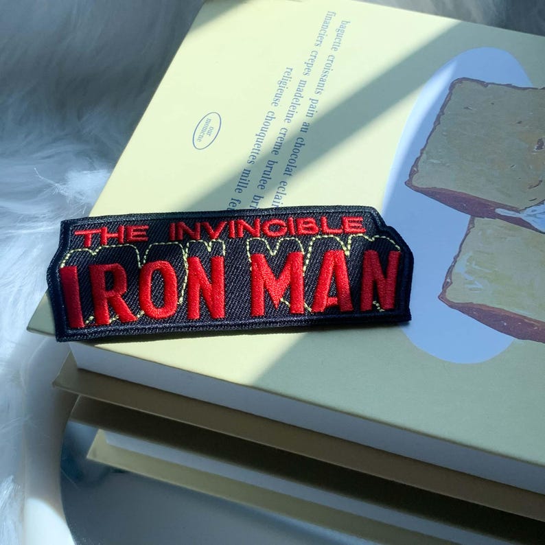 Puede incluir: Un parche bordado rectangular con fondo negro y letras rojas que dicen THE INVINCIBLE IRON MAN. El texto est&aacute; delineado con costuras amarillas y el parche presenta un dise&ntilde;o de tela estructurado con bordes limpios y definidos.