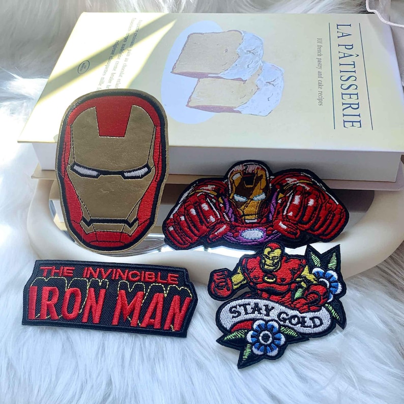 Puede incluir: Cuatro parches bordados con im&aacute;genes de Iron Man. Uno es un casco rojo y dorado, otro muestra a Iron Man volando, uno es un parche rectangular con el texto THE INVINCIBLE IRON MAN y otro muestra a Iron Man con flores y el texto STAY GOLD.