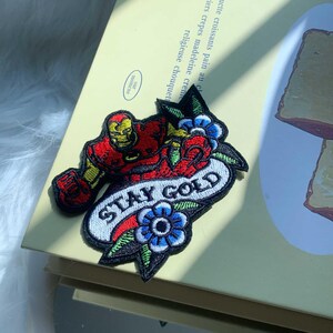 Puede incluir: Un parche bordado que presenta una figura con armadura roja y dorada, flores azules con hojas verdes y una pancarta blanca con el texto negro STAY GOLD. El dise&ntilde;o est&aacute; delineado con hilo negro y presenta un patr&oacute;n de puntadas detallado y colorido.