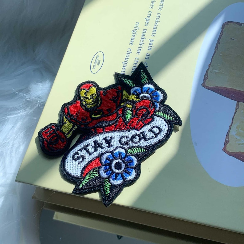 Puede incluir: Un parche bordado que presenta una figura con armadura roja y dorada, flores azules con hojas verdes y una pancarta blanca con el texto negro STAY GOLD. El dise&ntilde;o est&aacute; delineado con hilo negro y presenta un patr&oacute;n de puntadas detallado y colorido.