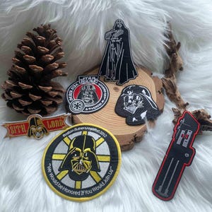 Puede incluir: Una colección de seis parches bordados de Star Wars con imágenes de Darth Vader. Los parches incluyen una silueta de cuerpo completo, un emblema circular de la Estrella de la Muerte, un perfil de casco, un estandarte de Sith Lord, un parche de Boston Super Megafest 2012 y un sable de luz.