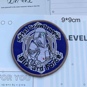 May include: A circular embroidered patch featuring a white Snowtrooper figure on a blue background. The text 501st Snowtrooper Blizzard Force is stitched in black around the figure. The patch has a tan border and measures 9 by 9 centimeters.
