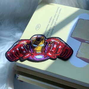 Puede incluir: Un parche bordado que muestra a un personaje de Iron Man estilizado con un traje rojo y dorado con los pu&ntilde;os extendidos, un reactor arc azul brillante en el pecho y un borde negro, colocado sobre una cubierta de libro amarillo claro con texto.