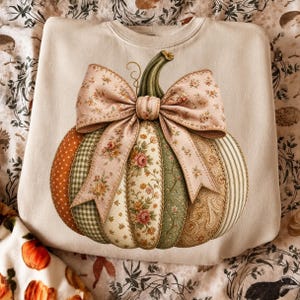 May include: A cream-colored crewneck sweatshirt featuring a large, central graphic of a pumpkin. The pumpkin is composed of various fabric-like segments in orange, green, and beige with floral, gingham, and paisley patterns, topped with a large bow.
