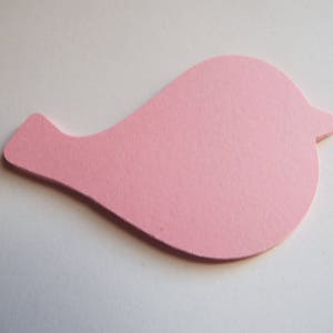 Op de afbeelding: Een platte, gestanste papieren vorm in de gedaante van een gestileerde vogel. De uitsparing is effen lichtroze met een gladde, matte textuur. Het vogel-silhouet heeft een rond lichaam, een puntige snavel aan de rechterkant en een kleine staart.