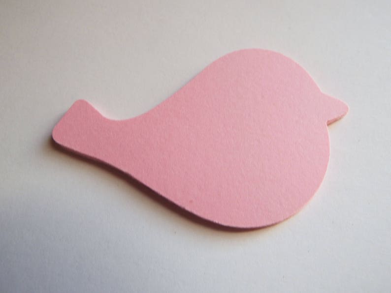Op de afbeelding: Een platte, gestanste papieren vorm in de gedaante van een gestileerde vogel. De uitsparing is effen lichtroze met een gladde, matte textuur. Het vogel-silhouet heeft een rond lichaam, een puntige snavel aan de rechterkant en een kleine staart.