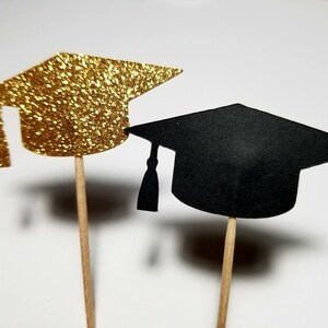 May include: Two graduation caps on wooden sticks. One cap is gold glitter and the other is black.
