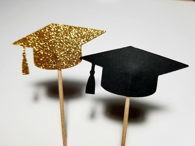 May include: Two graduation caps on wooden sticks. One cap is gold glitter and the other is black.
