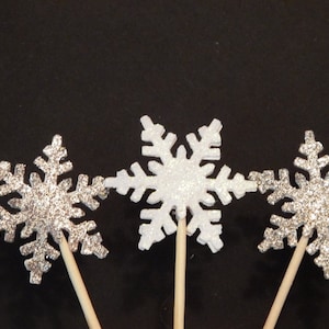 Könnte beinhalten: Drei silberne Glitzer-Schneeflocken-Kuchentopper auf Holzstielen. Die Schneeflocken sind unterschiedlich groß und haben einen weißen Hintergrund.