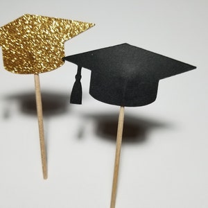 May include: Two graduation cap cupcake toppers, one gold glitter and one black. The toppers are attached to wooden sticks.