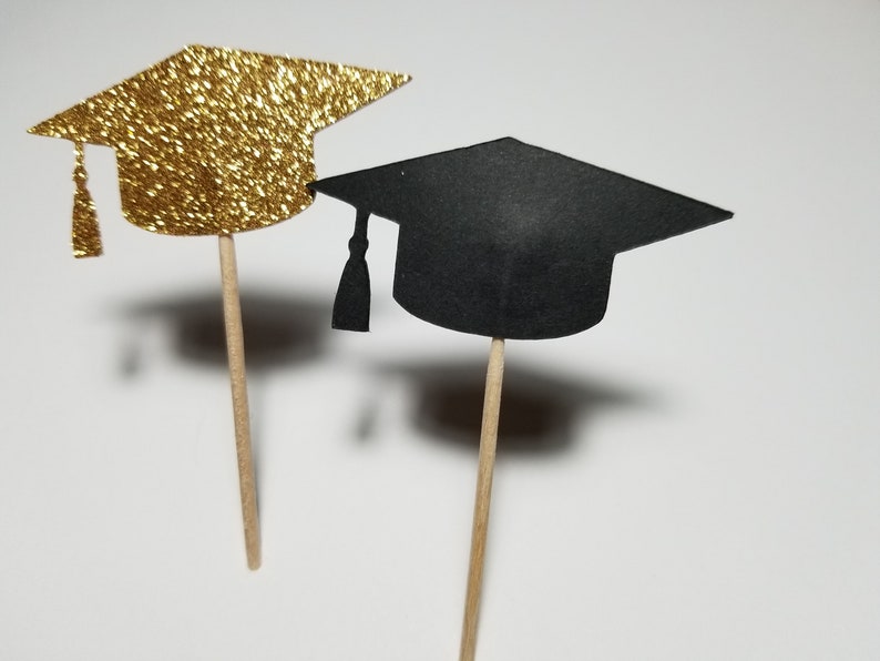 May include: Two graduation cap cupcake toppers, one gold glitter and one black. The toppers are attached to wooden sticks.