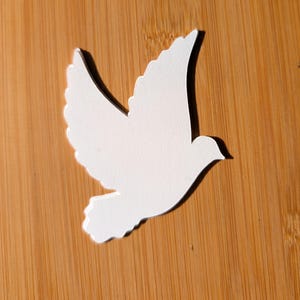 Peut inclure: Découpe de colombe en papier blanc avec des plumes d'ailes et de queue détaillées. La colombe est posée sur une surface en bambou brun clair, projetant une ombre. L'image est bien éclairée, soulignant la forme et la texture de la colombe.