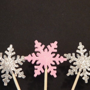 May include: Three snowflake cake toppers, one pink and two silver, with glitter accents. The snowflakes are attached to wooden sticks.