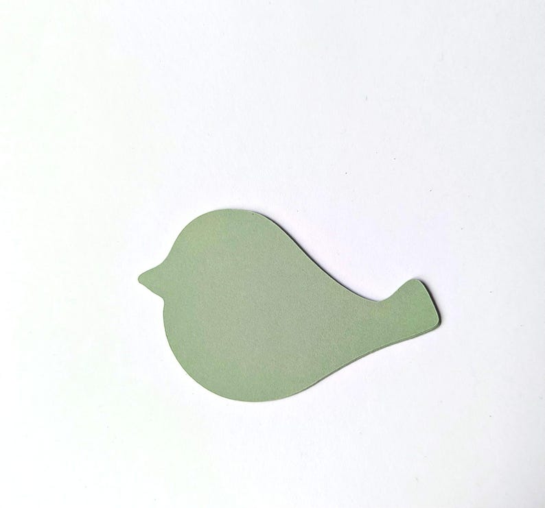 Op de afbeelding: Een platte, uitgestanste papieren vorm in de gedaante van een gestileerde vogel. Het vogel-silhouet is uitgevoerd in een effen, gedempte saliegroene kleur met een rond lichaam, een kleine puntige snavel en een korte, schuin aflopende staart.