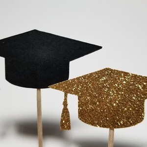 May include: Two graduation caps on sticks, one black and one gold glitter. The caps are perfect for decorating cupcakes or other treats for a graduation party.