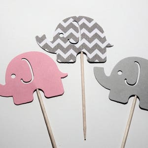 May include: Three elephant cupcake toppers, one pink, one gray with a chevron pattern, and one gray. Each topper is attached to a wooden stick.