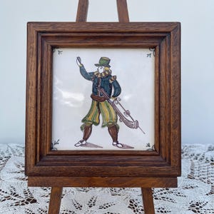 Puede incluir: Un azulejo de cerámica cuadrado con una ilustración pintada a mano de una figura con sombrero, chaqueta oscura y pantalones a rayas que sostiene un rifle largo. El azulejo está en un marco de madera marrón oscuro sobre un pequeño caballete.