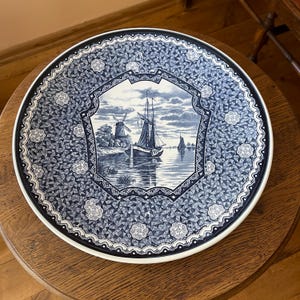 Può includere: Un piatto rotondo in ceramica con un motivo blu e bianco. Il centro raffigura una barca a vela sull'acqua vicino a un mulino a vento. Il bordo presenta un design geometrico a mosaico con piccoli motivi floreali e un bordo smerlato.