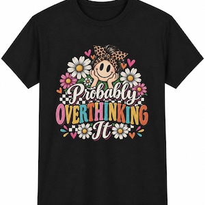 May include: A black t-shirt featuring a graphic print of a smiley face wearing a leopard print headband, surrounded by white and pink daisies, small hearts, and a checkered pattern. The text reads "Probably OVERTHINKING It" in colourful letters.