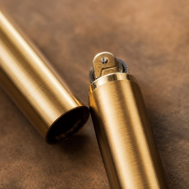 May include: A cylindrical brass lighter with a brushed metal finish, shown with its matching cap removed. The lighter features a visible ignition wheel and a small screw mechanism at the top, displaying a warm, metallic gold-toned surface texture.