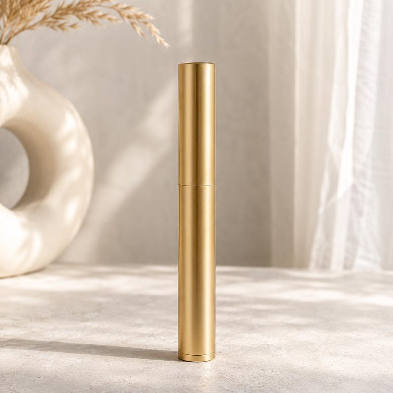 May include: A tall, cylindrical, metallic gold tube with a smooth, matte finish. The object features a clean, vertical design with a subtle horizontal seam dividing the upper cap section from the lower base, standing upright on a flat surface.