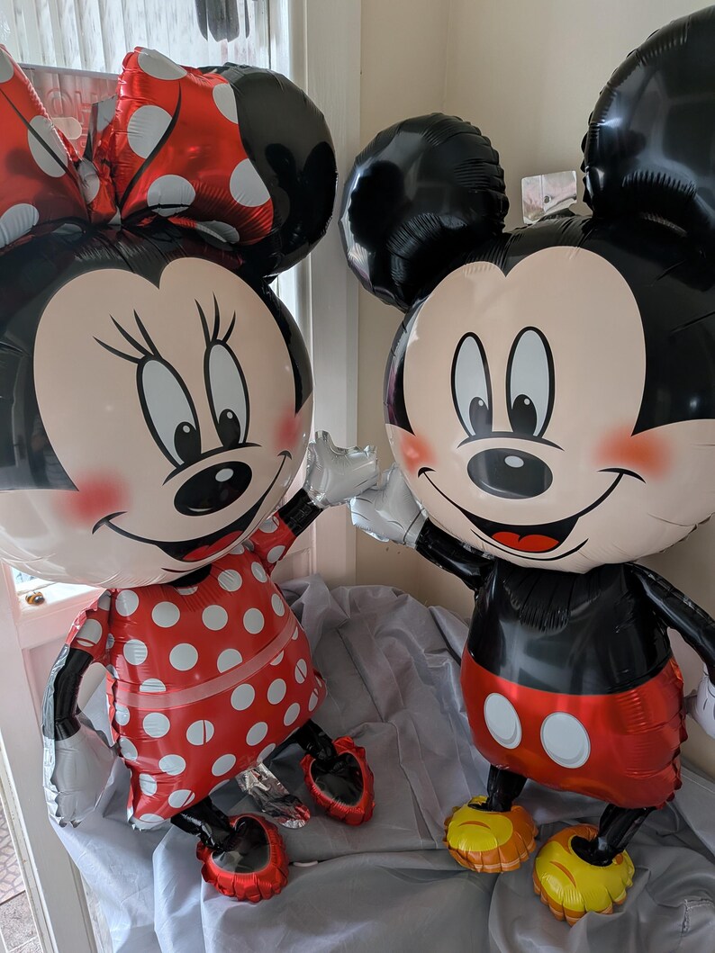 Może przedstawiać: Dwa balony foliowe w kształcie postaci z kresk&oacute;wek. Jeden balon ma czerwoną sukienkę w białe kropki i pasującą kokardę. Drugi balon ma czarne spodenki z dwoma białymi guzikami i ż&oacute;łte buty. Oba balony mają czarne uszy.