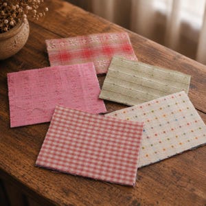 May include: Five rectangular fabric swatches featuring various patterns and textures. One is pink with small embroidered flowers, one is red plaid with floral borders, one is olive green with horizontal lines, one is red gingham, and one is cream with dots.