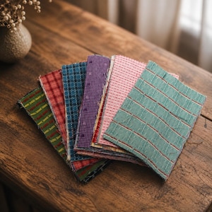 May include: A fan-shaped arrangement of rectangular fabric swatches featuring various patterns and colors, including green with yellow lines, red plaid, blue plaid, purple gingham, pink with dashed lines, and a light teal with horizontal stripes.