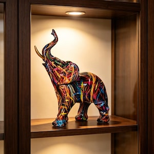 May include: A standing elephant figurine featuring a black base color with vibrant, multi-colored abstract paint streaks in red, yellow, blue, and white. The elephant has its trunk raised upward and features a single prominent gold-colored tusk.